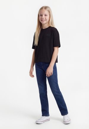 Young girl with long blonde hair wearing a black shirt, blue jeans, and white sneakers, standing and smiling against a white background.