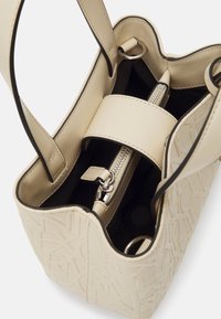 Kremowa skórzana torba typu tote z tłoczonymi geometrycznymi wzorami, górną komorą zapinaną na suwak oraz srebrnymi akcentami metalowymi. Otwarta góra z podwójnymi uchwytami.