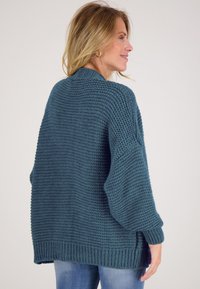 Teal gebreide trui met een gestructureerd patroon, verlaagde schouders en geribbelde mouwen. Ontspannen pasvorm met een langere achterkant.