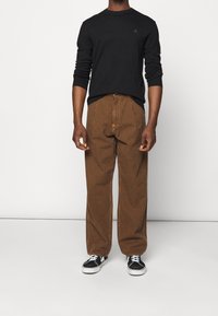 Sudadera negra, pantalones anchos a rayas marrones y zapatillas negras con suelas blancas. El conjunto presenta un pequeño logo en la sudadera.