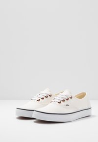 Leinensneaker mit cremefarbener Außenseite, weißen Schnürsenkeln, bronzefarbenen Ösen und einem schwarzen Sohlenakzent. Rundliche Zehenpartie und flaches Design.