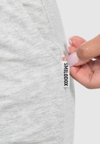 Shorts jogger grigio chiaro realizzati in tessuto morbido e testurizzato, con un'etichetta del marchio ben visibile. Una mano sta tirando l'etichetta vicino a una tasca laterale.
