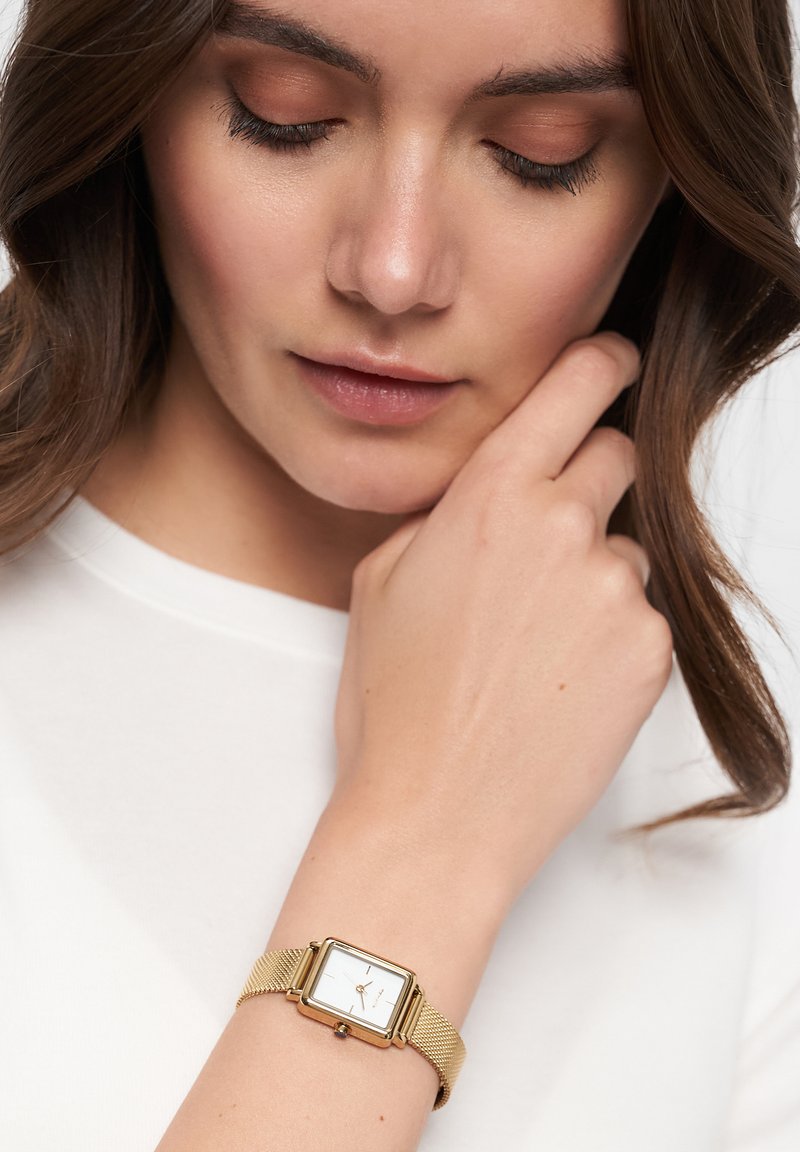 Montre avec un bracelet en maille dorée et un cadran carré blanc, dotée de marqueurs d'heures minimalistes et d'une petite couronne. Positionnement sur le poignet visible.