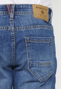 Los jeans de mezclilla azul oscuro presentan un corte recto, bolsillos traseros con costuras amarillas y una etiqueta marrón con el logo de la marca en la cintura.