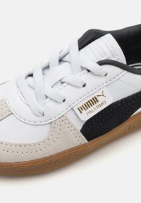 Puma Palermo sneakers har en hvit lær overdel med svarte og lysegrå semskede detaljer, flate hvite snøringer og en brun gummisåle.