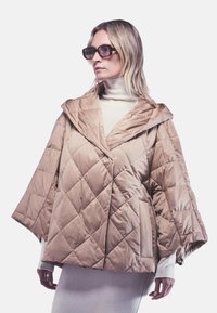 Teddy jacket in tan met diamantpatroon, oversized capuchon en brede mouwen. Draagt over een witte hoge hals top met een gladde textuur.