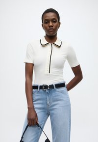 Λευκό ριμπ τοπ με μαύρο γείσο και γιακά, με κουμπιά στο μπροστινό μέρος. Συνδυασμένο με ανοιχτό μπλε ψηλόμεσα τζιν και μαύρη ζώνη.