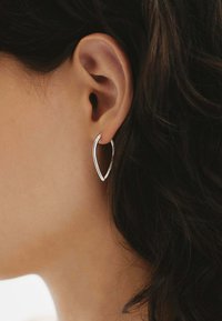 Pendientes de aro plateados con forma de lágrima, que presentan un acabado suave y pulido, y un diseño abierto, llevados en la oreja.