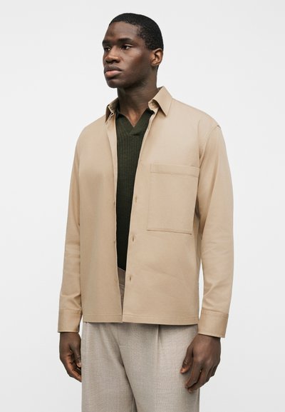 Άνδρας που φοράει μπεζ πουκάμισο με κουμπιά πάνω από σκούρο πράσινο πουλόβερ με V λαιμόκοψη και ανοιχτό γκρι παντελόνι, στέκεται μπροστά σε ένα απλό λευκό φόντο.