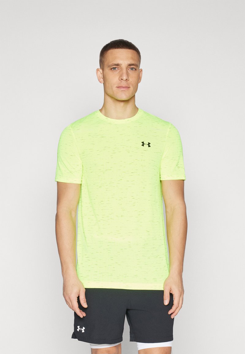Neon žluté sportovní tričko s krátkým rukávem a texturovaným vzorem, s černým logem na hrudi. Slačeno s tmavými šortkami.