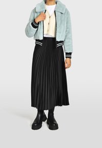 Bomber jacket in pelliccia sintetica azzurro chiaro con polsini a righe bianco e nero, indossato sopra un cardigan beige e una gonna plissettata nera, abbinato a robusti stivali neri.