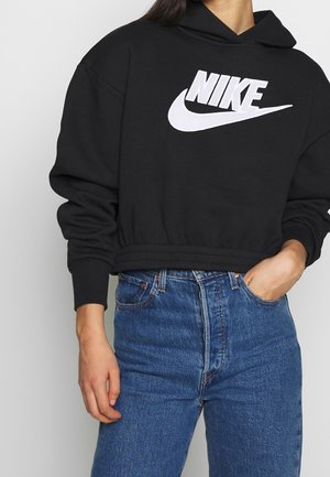 Persoon draagt een zwarte Nike-hoodie met een groot wit logo en blauwe jeans met een hoge taille, één hand in de zak, neutrale achtergrond.