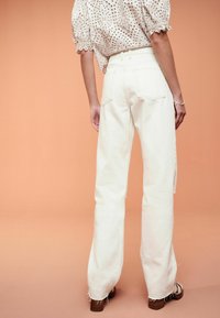 Jeans bianchi a vita alta con vestibilità dritta, orli sfrangiati e tasche posteriori, abbinati a una blusa avorio con motivo.