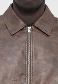 Veste marron en cuir synthétique avec un col à revers, dotée d'une fermeture éclair centrale en argent et d'une surface texturée.