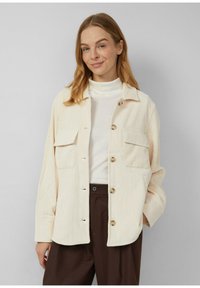 Crème kleurige corduroy shirtjas met twee borstzakken, knoopsluiting en een ontspannen pasvorm. Gedragen over een witte turtleneck en een paarse broek.