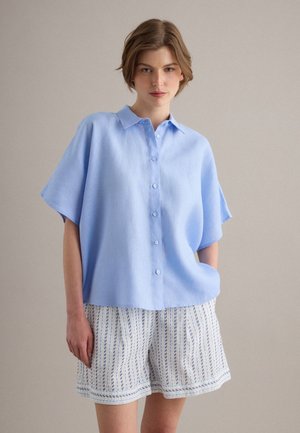 Giovane con una camicia azzurro chiaro ampia con bottoni e pantaloncini bianchi con righe verticali blu, in piedi davanti a uno sfondo semplice.