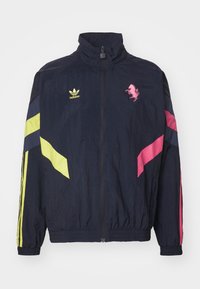 Marine blå vindjakke med høj krave, lynlås lukning og farverige detaljer i gul og pink. Inkluderer Adidas-logo og et pink grafik.