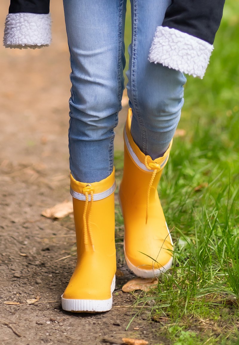 Bottes de pluie en caoutchouc jaune avec des accents blancs, finition brillante et détail de fermeture éclair, portées avec un jean bleu dans un cadre naturel en plein air.