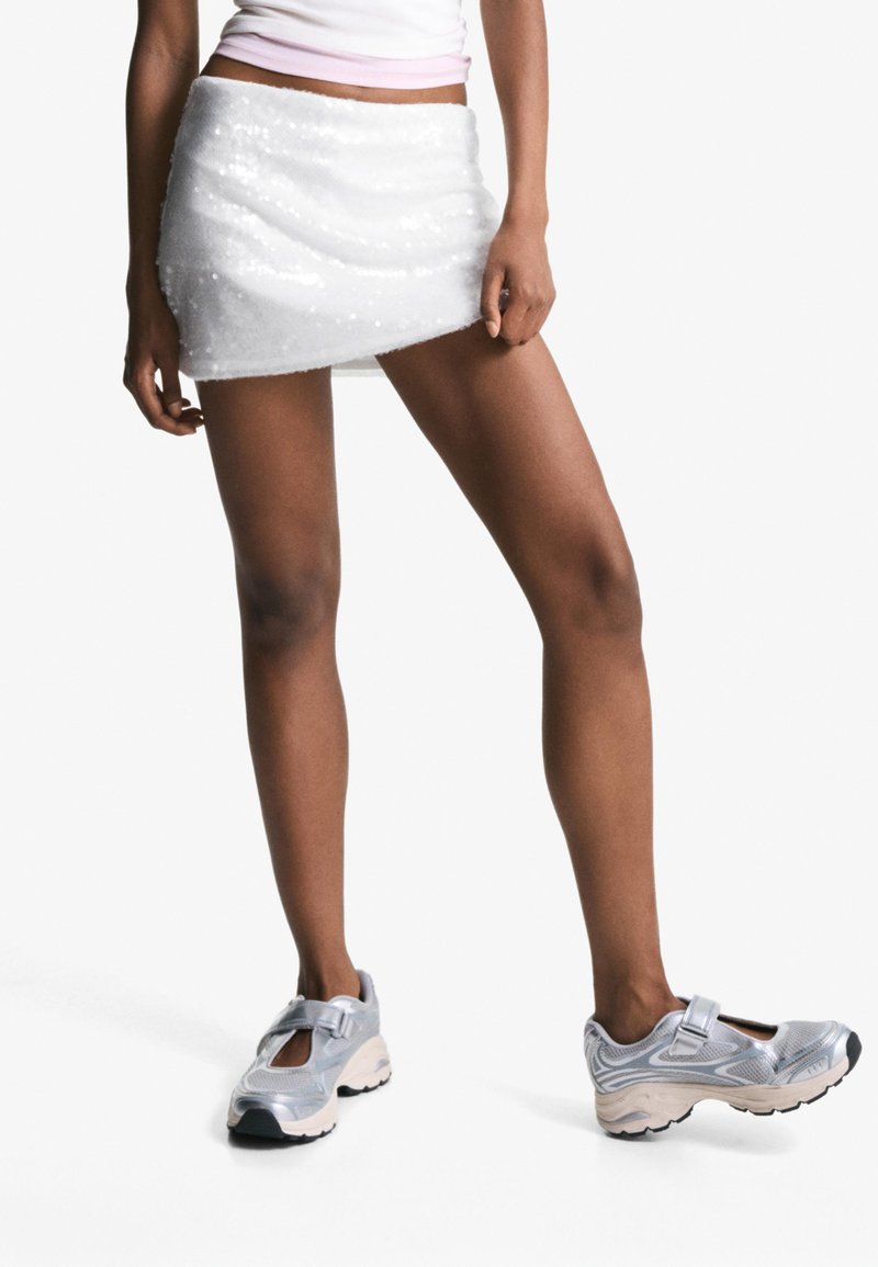 Femeie îmbrăcată cu o fustă mini albă cu paiete, un top alb scurt și adidași sportivii argintii, stând pe un fundal alb uniform.