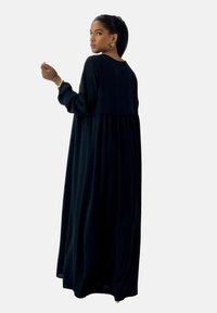 Vestido negro de manga larga con un corte relajado, que cuenta con una cintura fruncida y una textura suave, complementado con pendientes dorados y un peinado de coleta.