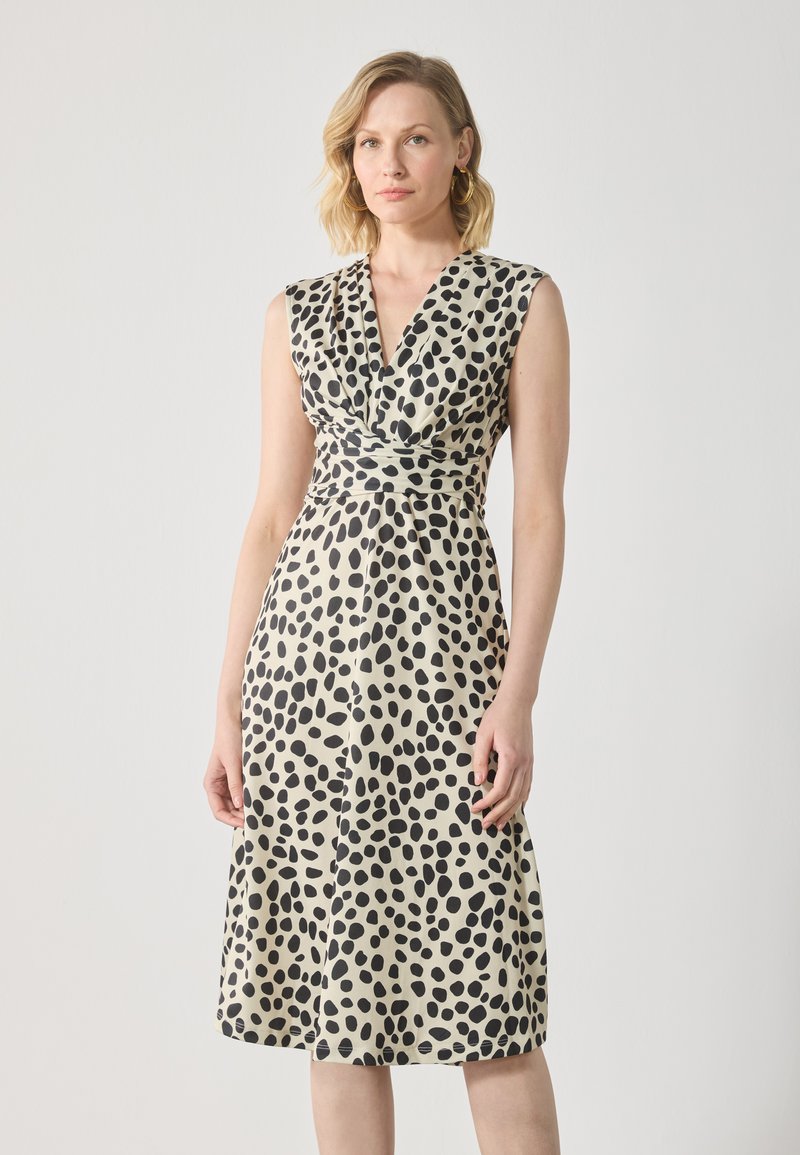 Robe mi-longue en tissu crème avec un motif de points noirs irréguliers, présentant un décolleté en V profond et un buste en style cache-cœur. Sans manches.