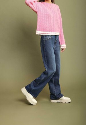 Personne portant un pull rose texturé, un jean bleu ample et des bottes blanches épaisses, debout devant un fond olive uni.