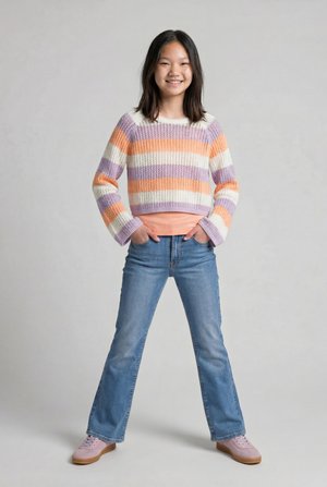 Jong persoon staat met handen in de zakken, draagt een gestreepte pasteltrui, blauwe jeans en roze sneakers, glimlachend tegen een effen achtergrond.