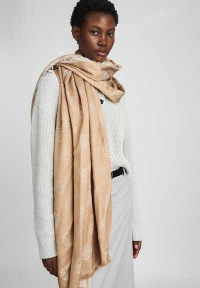 Beige κασκόλ με διακριτό μοτίβο λογοτύπου, με φούντες στην άκρη και απαλή υφή, απλωμένο πάνω σε ένα ανοιχτό γκρι πουλόβερ.