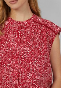 Rode blouse met een geometrisch wit patroon, korte kapmouwen, een knoopsluiting bij de hals en plooien voor extra textuur en volume.