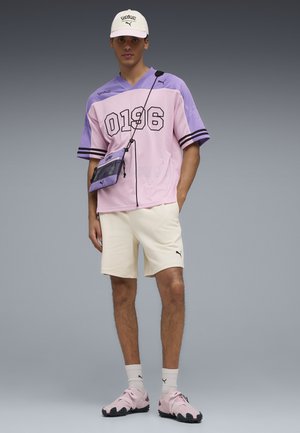 Jonge man met een sporttrui in pastelpaars, beige korte broek, roze sneakers, witte sokken, een witte pet en een schoudertasje.