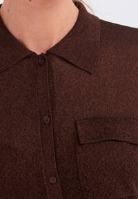 Brun skjorte med knapper og en struktureret overflade. Har en krave, to frontknapper og en brystlomme. Klassisk design og varm farvetone.