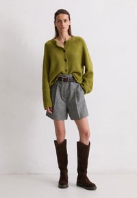 Cardigan verde lavorato a maglia con bottoni, pantaloni kaki grigi su misura e stivali marroni fino al ginocchio, il tutto su uno sfondo neutro.