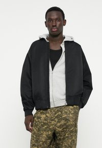 Črno bomber jakno z gladkim zaključkom, kombinirano s sivo kapucarjo z zadrgo in cargo hlačami z vzorcem kamuflaže. Enostavna zasnova, sproščen kroj.