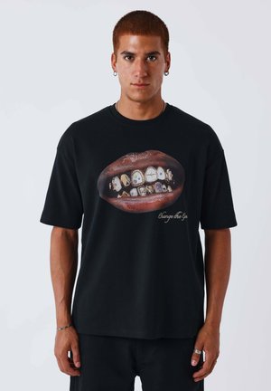 Uomo con capelli corti rossicci che indossa una maglietta nera con grandi labbra con denti decorati e la scritta "Change the Gen" sul davanti.