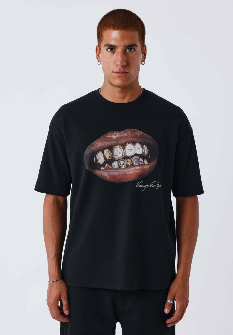 Homme aux cheveux courts rougeâtres portant un t-shirt noir avec une grande bouche aux dents décorées et le texte « Change the Gen » sur le devant.
