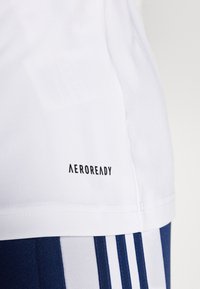 Camisa deportiva blanca con el logo "AEROREADY" en negro, con tejido suave y un dobladillo estructurado. Acentos de rayas en azul marino visibles en la parte inferior.
