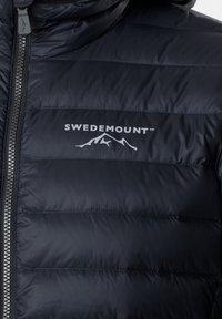 Veste matelassée noire avec fermeture éclair devant et logo Swedemount avec un motif montagne sur la poitrine.