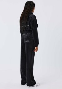 Zwart velours trainingspak met een cropped jack en wijde broek. Het jack heeft een hoge kraag; de broek heeft zakken en logoelementen.
