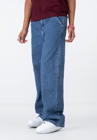 Jeans lejere de culoare albastră, confecționate din denim, având un design clasic cu cinci buzunare, cusături vizibile și o margine desfăcută. Purtați cu adidași albi.