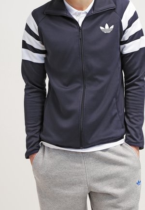 Homme portant une veste Adidas bleu marine zippée avec des bandes blanches sur les manches et un pantalon de jogging Adidas gris, mains dans les poches.