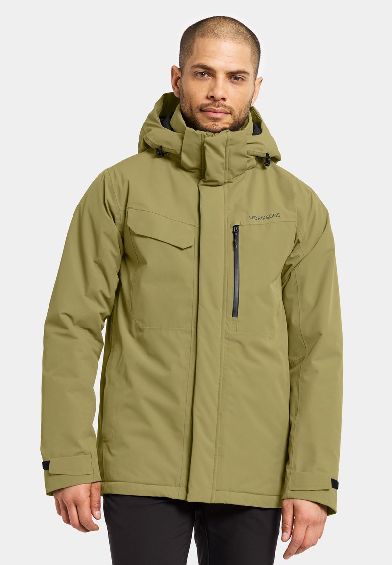 Homme portant une veste d'hiver à capuche de couleur vert olive avec une poche poitrine zippée et une poche poitrine à rabat, se tenant devant un fond uni.