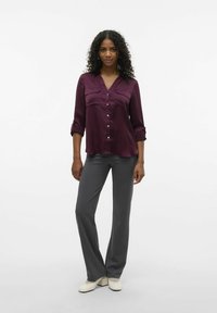 Blouse en satin bourgogne avec fermeture à boutons et deux poches poitrine, assortie à un pantalon à jambes larges gris et des chaussures à talons blocs couleur crème.