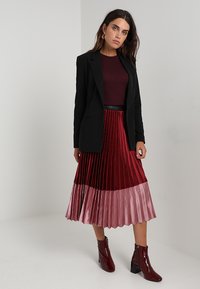 Sort blazer over en bourgogne top, parret med en plisseret nederdel med mørkerøde og lyserøde sektioner. Modellen bærer skinnende bourgogne ankelstøvler.