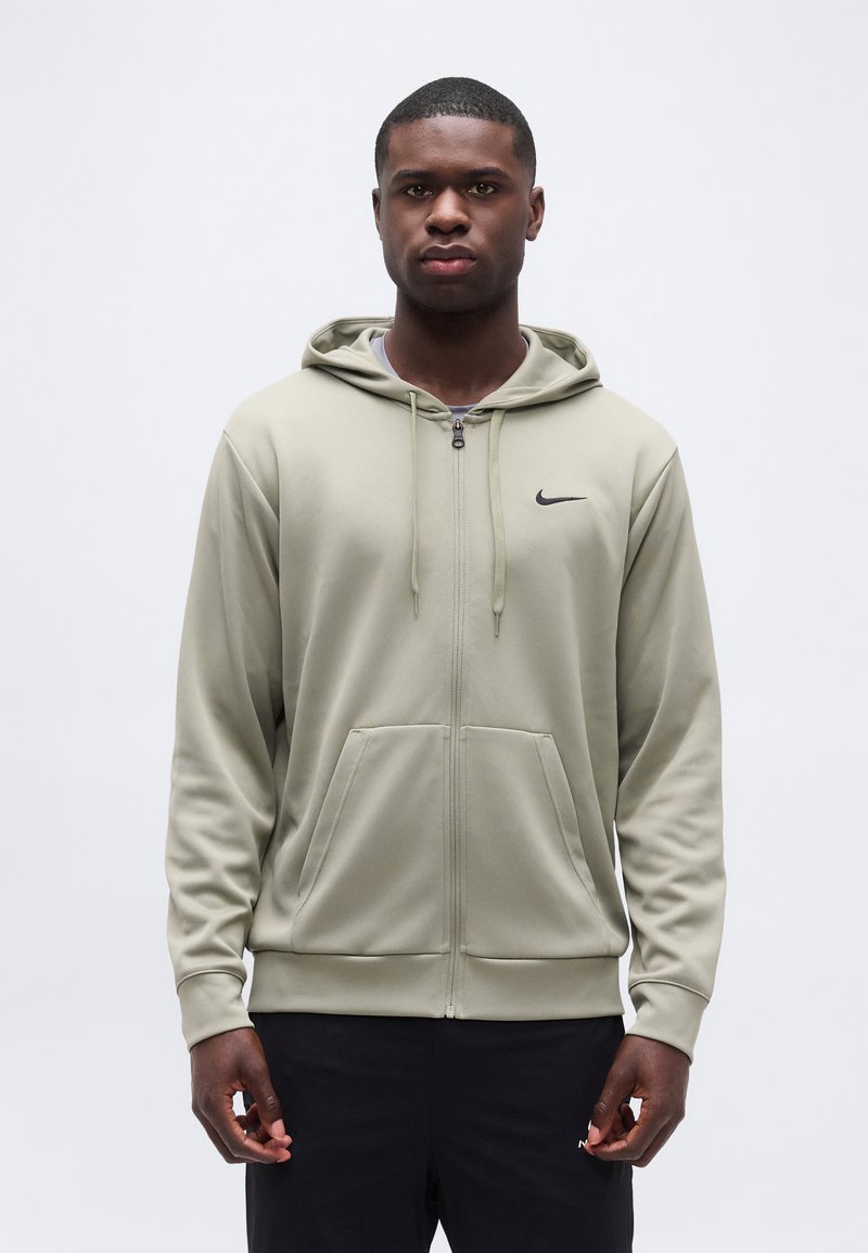 Άνδρας που φοράει ελαφρύ φωτεινό ελιά φούτερ Nike με φερμουάρ και μπροστινές τσέπες, καθώς και μαύρο αθλητικό παντελόνι, στέκεται μπροστά σε απλό λευκό φόντο.