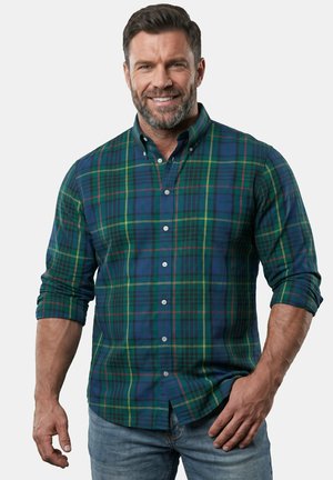 Ein Mann lächelt, trägt ein grün-blau kariertes Hemd mit Knöpfen und hochgekrempelten Ärmeln sowie hellblaue Jeans und steht vor einem einfarbigen Hintergrund.