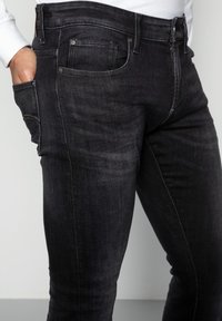 Jeans de mezclilla oscura con corte ajustado, que presentan un ligero efecto de desgaste, diseño de cinco bolsillos y costuras en contraste. Textura suave.