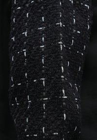 Μαύρο υφασμάτινο ύφασμα με υφή και σχέδιο πλέξης με κορδέλες που σχηματίζουν πλέγμα, με επαναλαμβανόμενο λευκό κείμενο "KARL LAGERFELD".