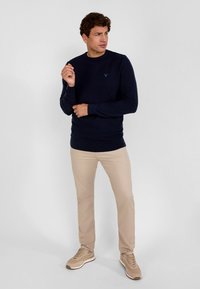 Jūrinio stiliaus apvalios iškirptės megztinis, pagamintas iš švelnios medžiagos, derinamas su šviesiai rudos spalvos liesomis kelnėmis ir šviesiai rudais sportbačiais, su subtiliu logotipo akcentu.