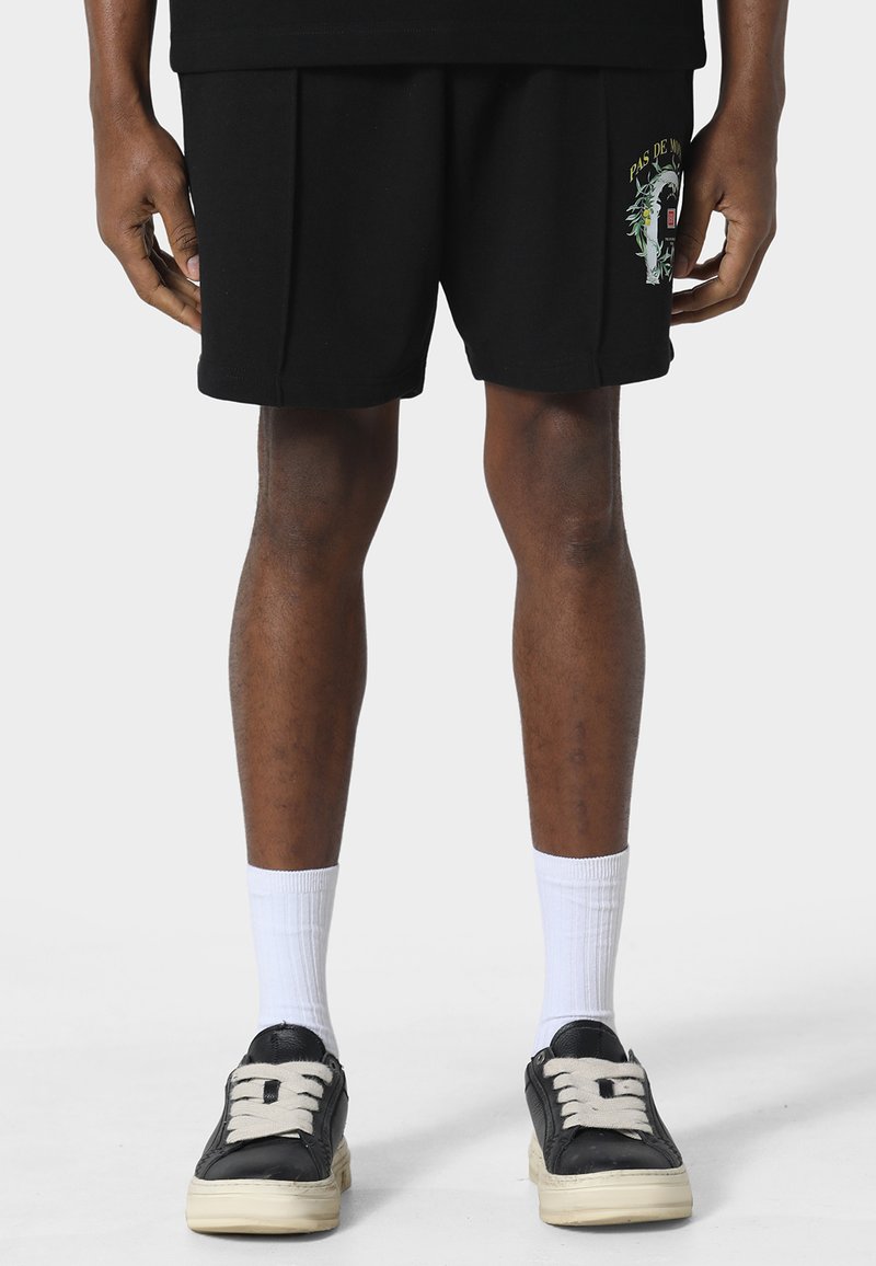 Jambes d'une personne portant un short noir avec un motif graphique, des chaussettes blanches et des baskets noires avec de gros lacets blancs, se tenant devant un fond uni.