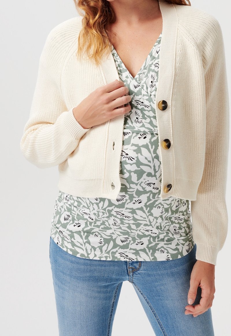 Femme portant un cardigan en maille crème avec des boutons en écaille de tortue sur un haut vert à fleurs en V, un jean bleu, main ajustant le cardigan.